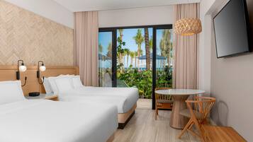A hotel room with two beds, a TV, a small table with chairs, and a view of palm trees and the sea.