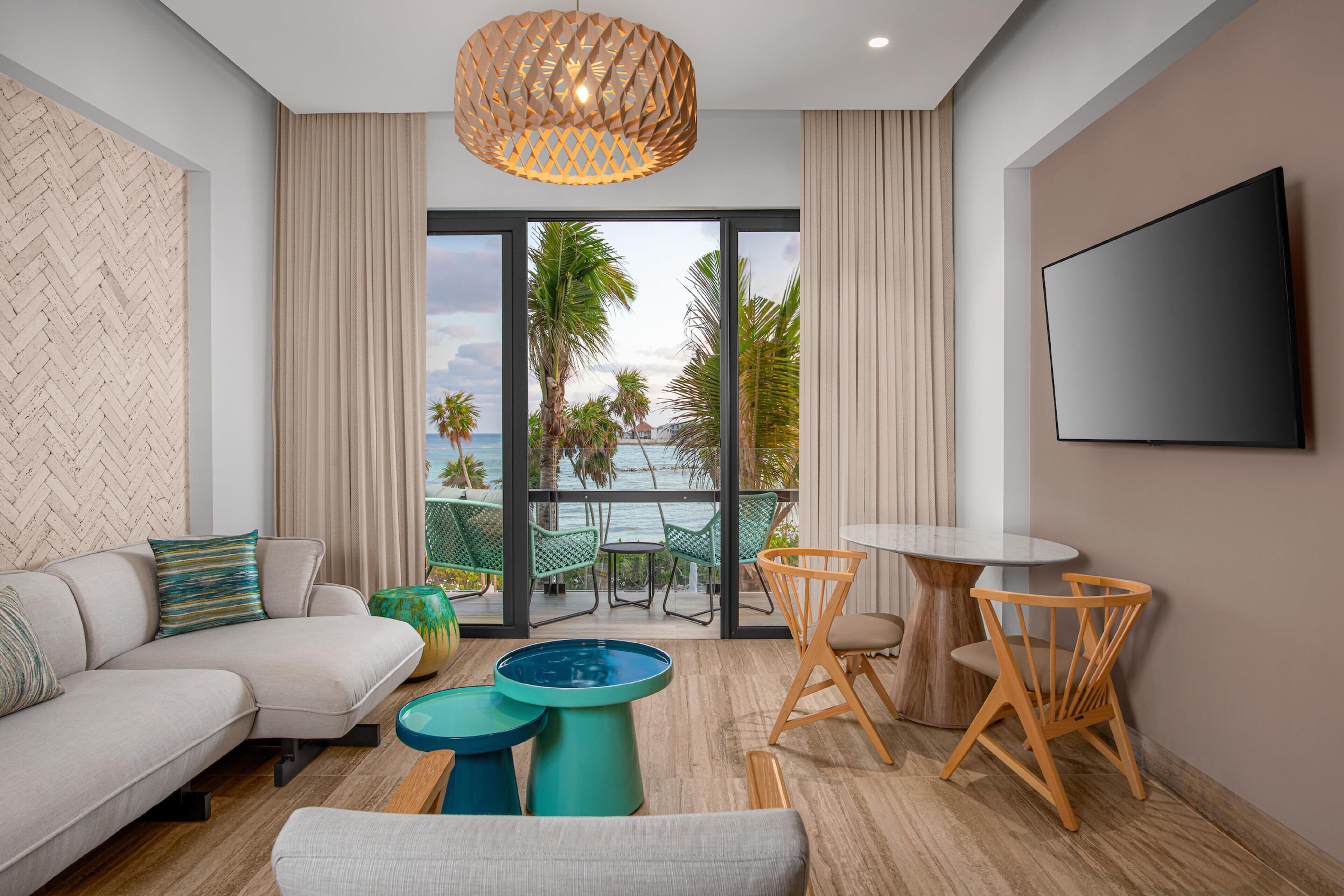 A modern living room with a sofa, coffee table, and chairs, a large TV, and a balcony with a view of palm trees and the sea.