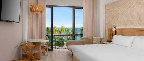 A hotel room with a large bed, a dining table with chairs, a TV mounted on the wall, and a view of the ocean through large windows.