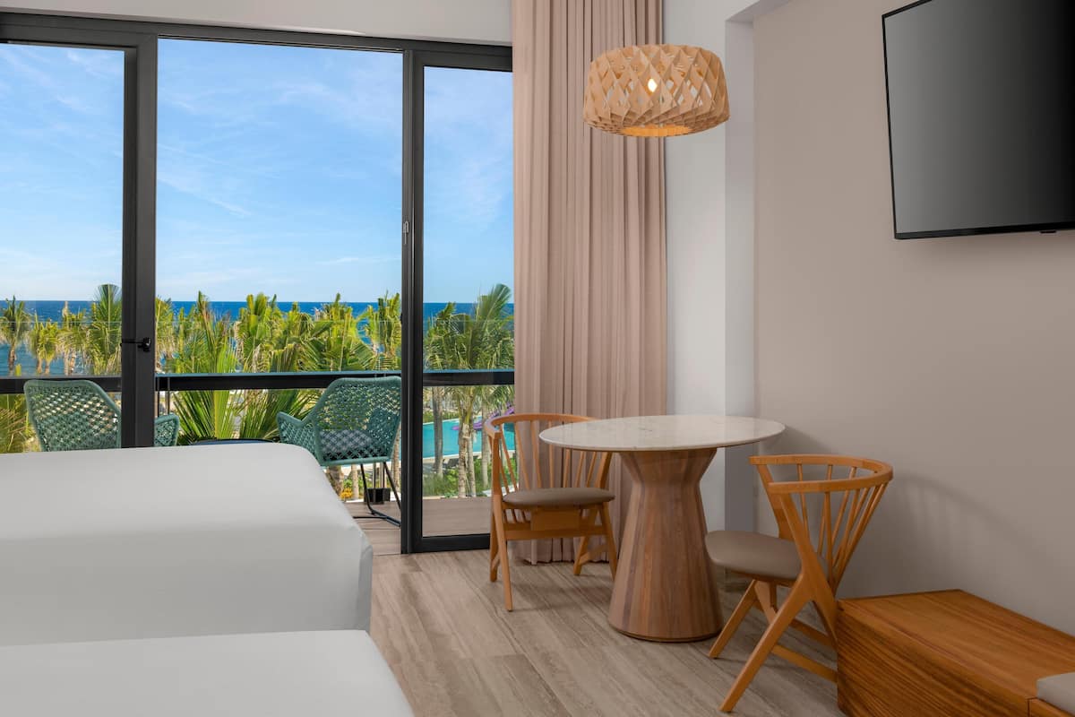 A hotel room with a balcony, a round wooden table, two wooden chairs, a TV mounted on the wall, and a large window offering a view of the ocean and palm trees.