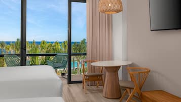 A hotel room with a balcony, a round wooden table, two wooden chairs, a TV mounted on the wall, and a large window offering a view of the ocean and palm trees.