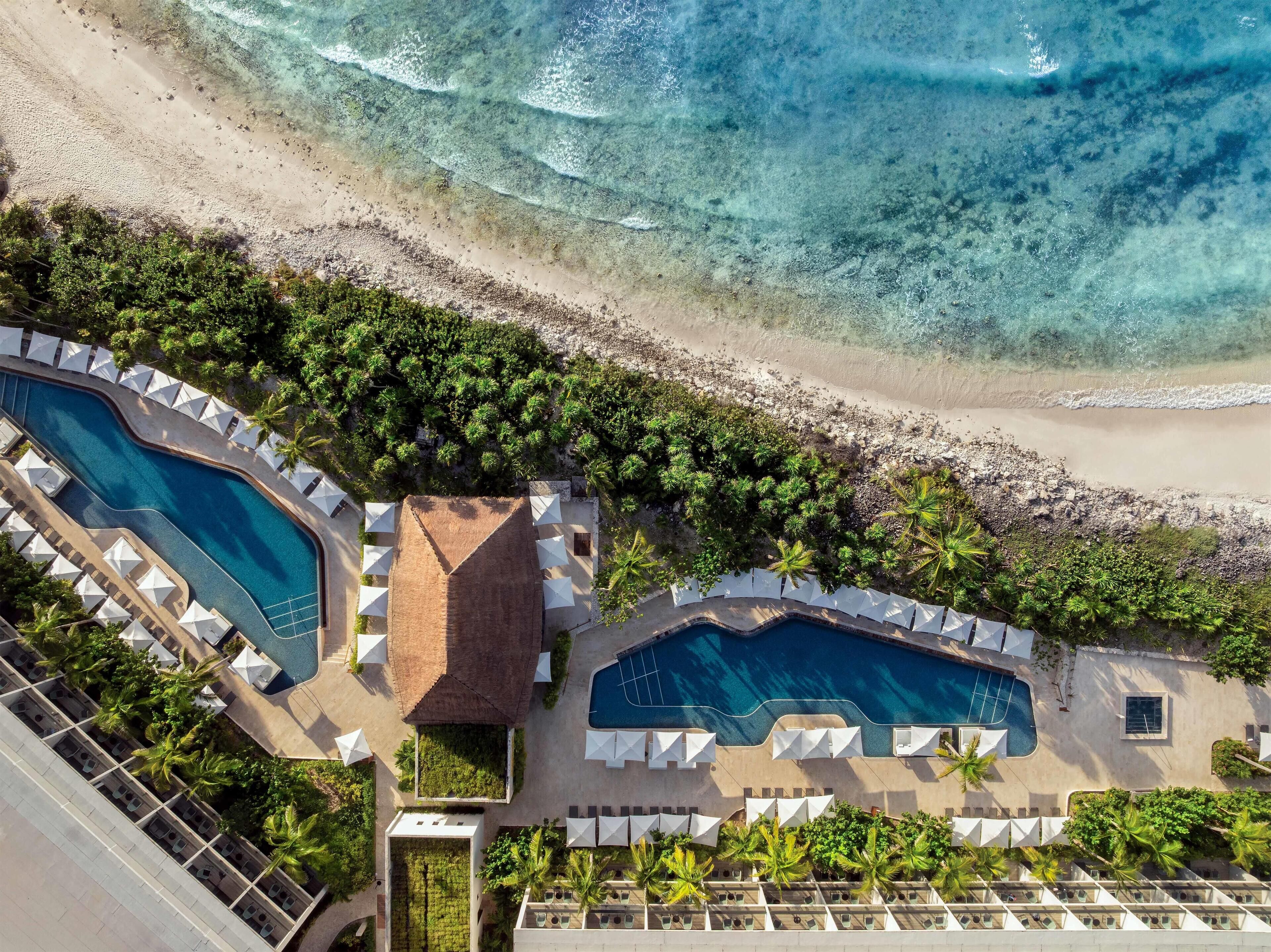Vista aérea de un resort con dos piscinas, una playa y abundante vegetación.