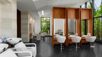 A modern salon with a clean, minimalist design, featuring white chairs, a wooden reception desk, and large windows offering a view of greenery.