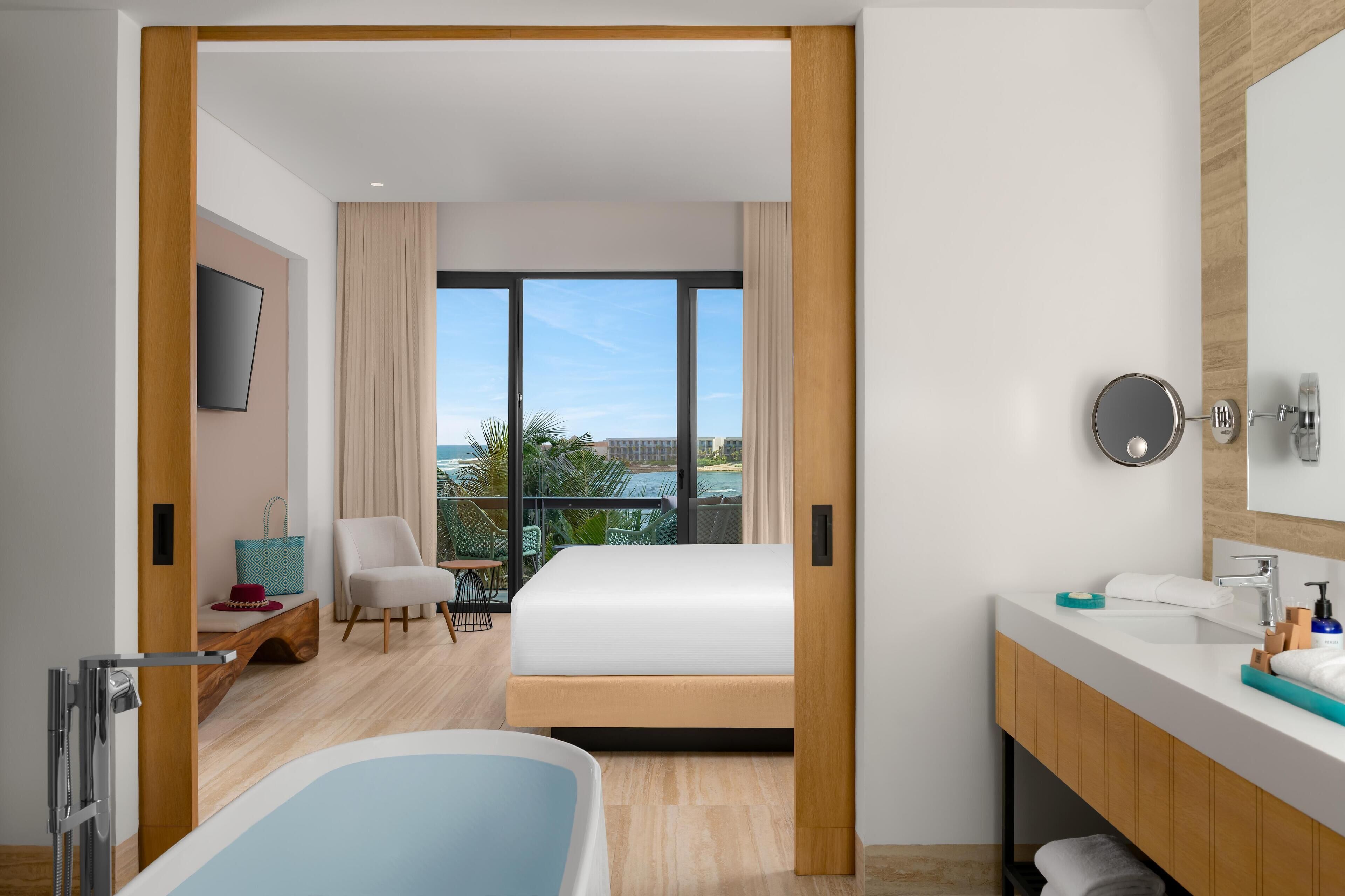 A modern hotel room with a large bed, a bathroom with a bathtub, and a balcony with a view of the city.