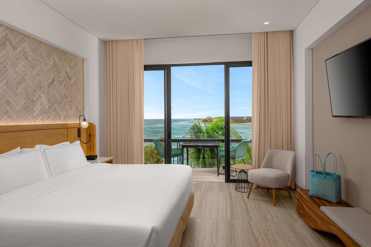 A hotel room with a large bed, a desk with a chair, a TV, and a balcony with a view of the ocean.