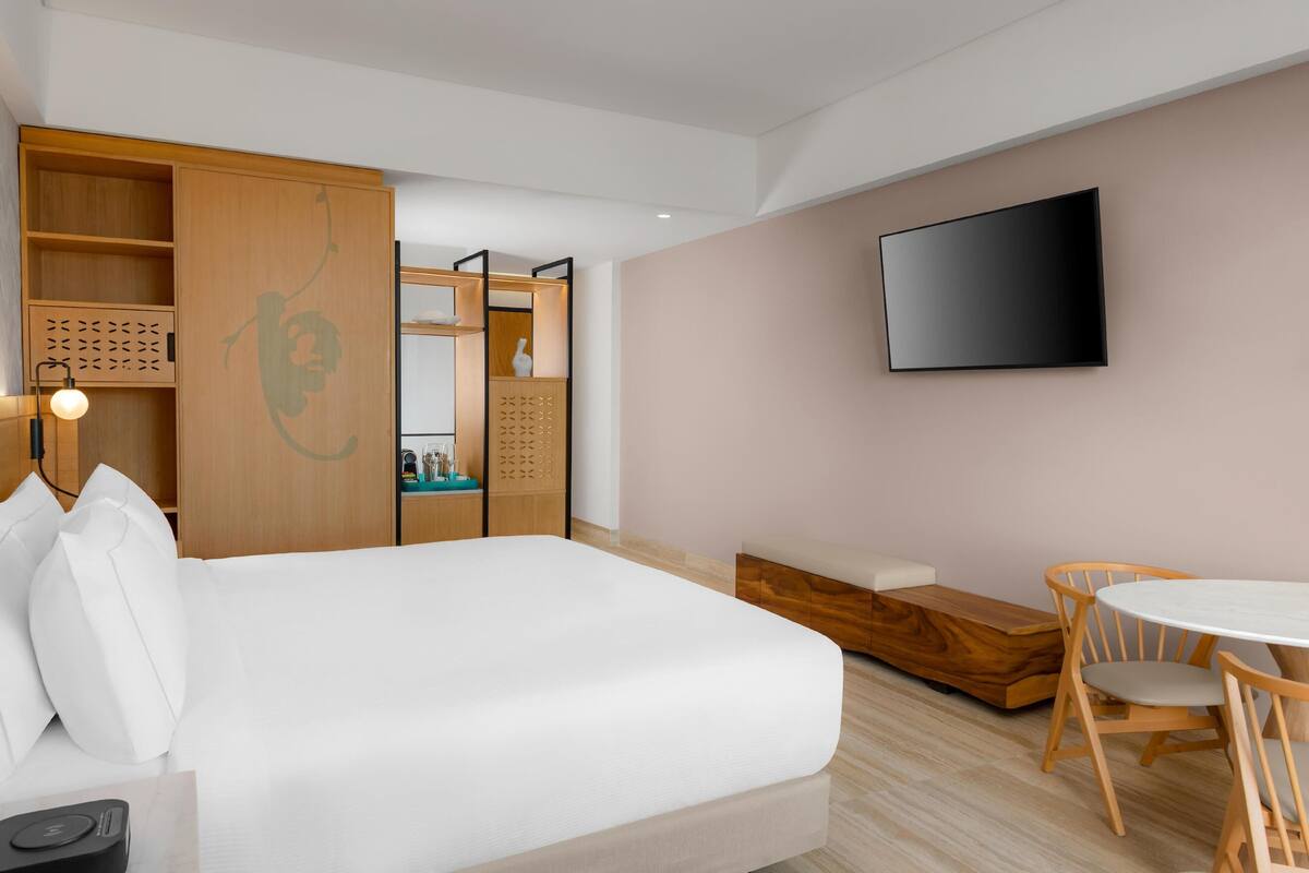 A modern hotel room with a bed, a TV, a wooden wardrobe, a bedside table, and a seating area with chairs.