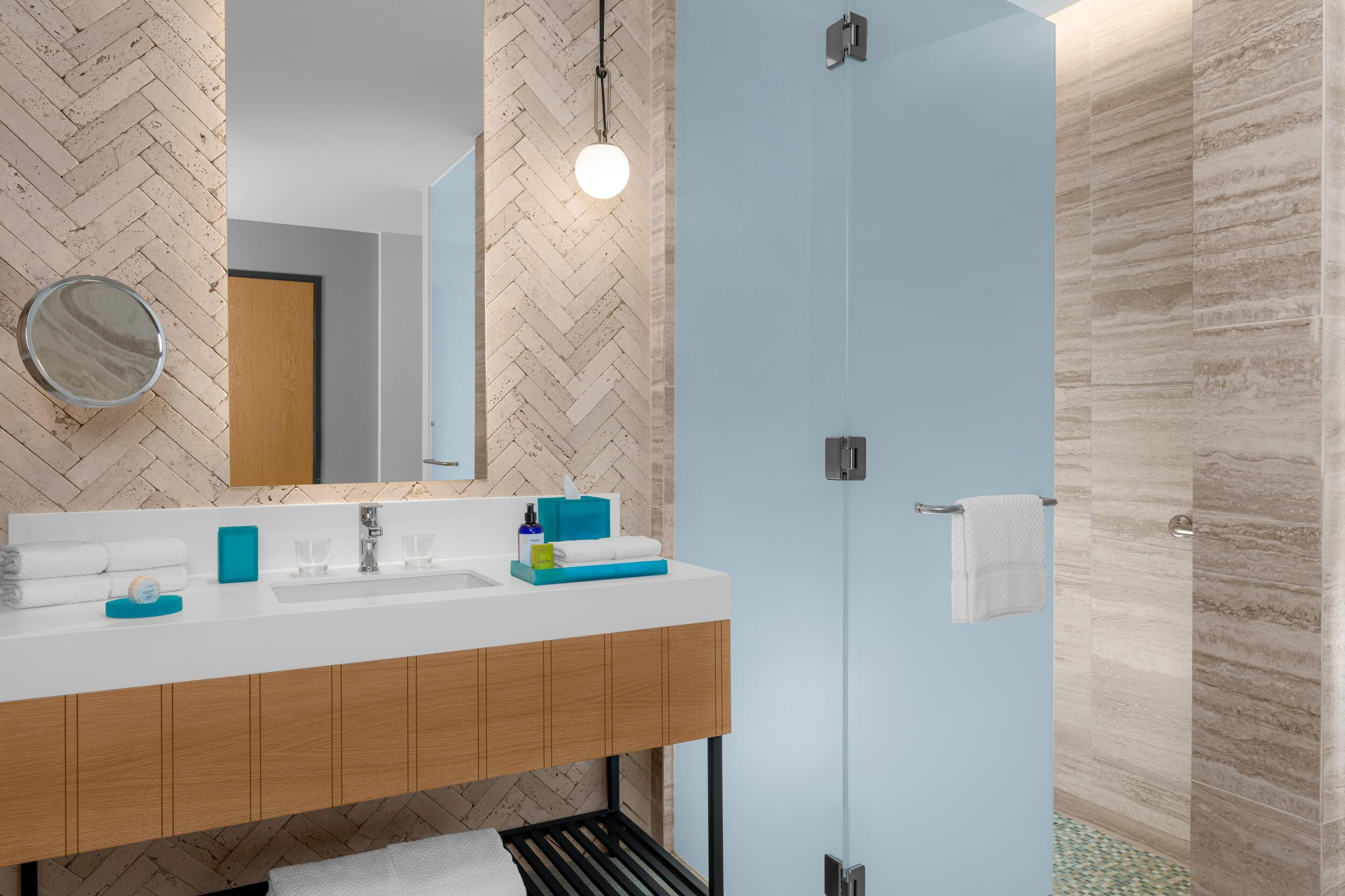 A modern bathroom with a herringbone-patterned wall, a white sink with a wooden cabinet below, a round mirror, and a glass shower enclosure.