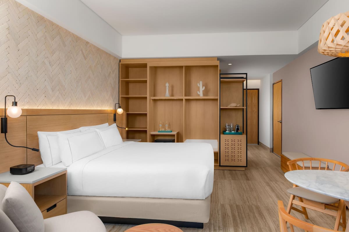 A modern hotel room with a large bed, wooden shelving, a desk, and a dining area.
