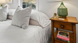 A bedroom with a bed, a nightstand with a lamp, a window, and a wall with a picture.