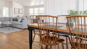 A modern living room with a grey sofa, wooden dining table, and chairs, a wooden floor, and a white door in the background.