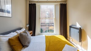 A hotel room with a bed, a window with curtains, a radiator, and a framed picture on the wall. - Ingram Apartments (Glasgow)