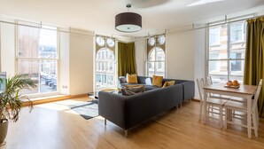 A modern living room with a sofa, dining table, and chairs. Large windows offer a view of the city. - Ingram Apartments (Glasgow)