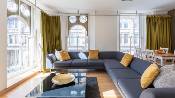 A modern living room with a grey sectional sofa, green curtains, and a dining area with a table and chairs. - Ingram Apartments (Glasgow)