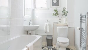 Un bagno moderno con vasca, lavabo, wc e un piccolo cestino. Sono presenti due finestre che illuminano naturalmente l'ambiente e un quadro appeso alla parete.