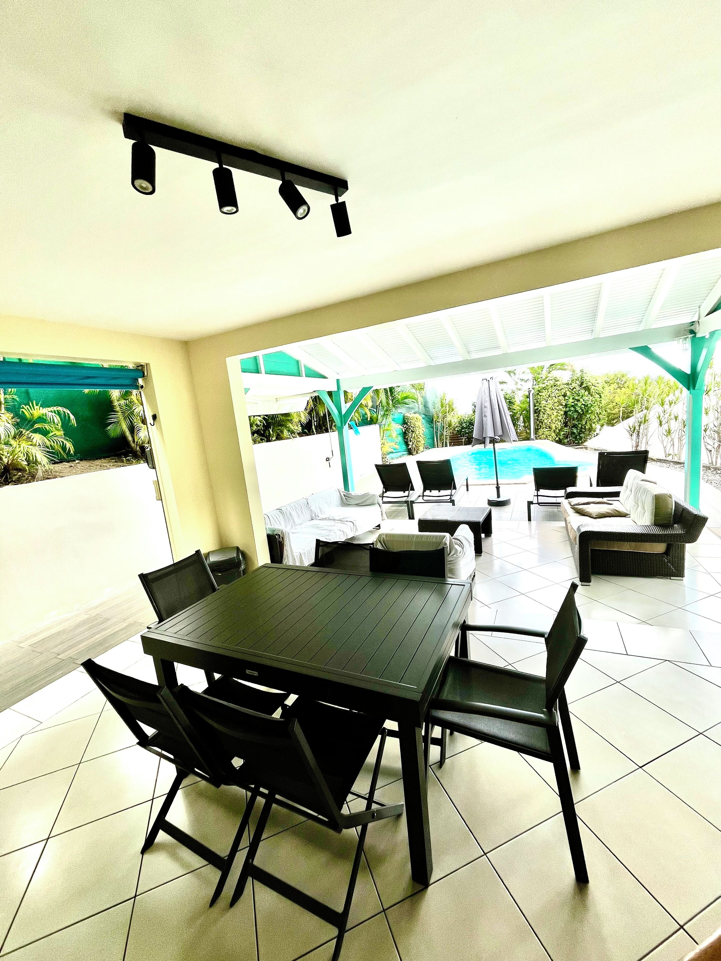 An outdoor patio area with a dining table and chairs, a pool in the background, and a covered seating area.