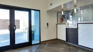 Vestíbulo de hotel con puerta corredera de vidrio, mostrador de recepción y piso de madera en patrón de espiga.