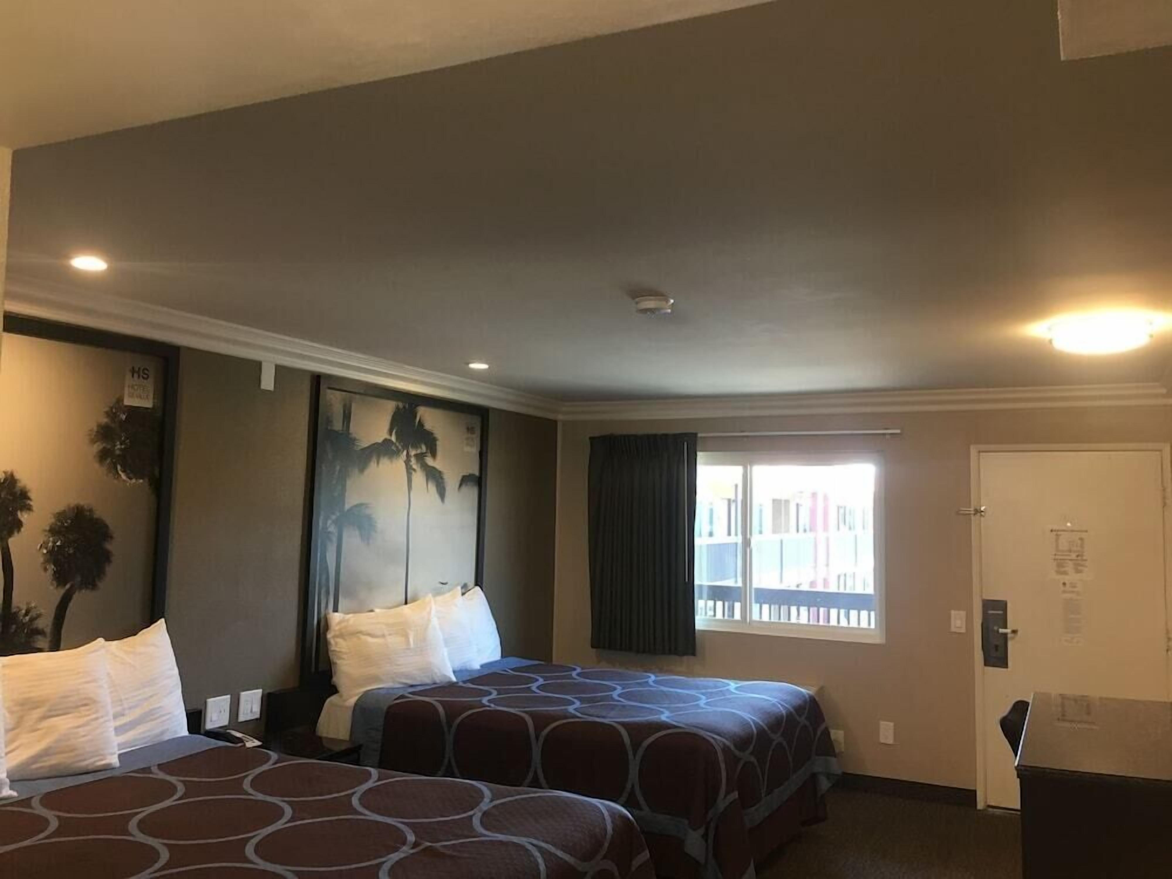 Habitación de hotel con dos camas, un ventanal con cortinas, una puerta con cerradura y un escritorio pequeño.