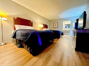 A hotel room with two beds, a wooden floor, a television on a dresser, and a nightstand with a lamp. - Howard Johnson Tulsa East/airport (Tulsa)