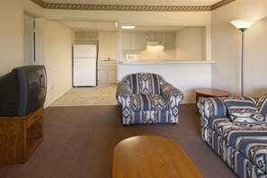 Una sala de estar con televisor, sillón estampado, sofá y una mesita. Se ve una cocina al fondo.