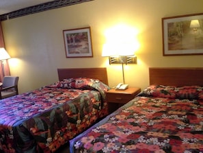 Una habitación de hotel con dos camas, cada una con ropa de cama floral, una mesita de noche con lámpara y un cuadro en la pared.