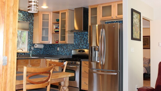A compact kitchen with wooden cabinets, a stainless steel refrigerator, and a dining area with a wooden table and chairs. - Serendipity House (Colorado Springs)