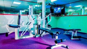 Ein Fitnessraum mit verschiedenen Trainingsgeräten, einem an der Wand montierten Fernseher und einem violetten Teppichboden.