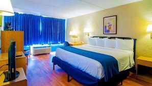 Ein Hotelzimmer mit einem großen Bett, einem Schreibtisch mit Computer, einem Sessel, einer Kaffeemaschine und einem Fenster mit Vorhängen.