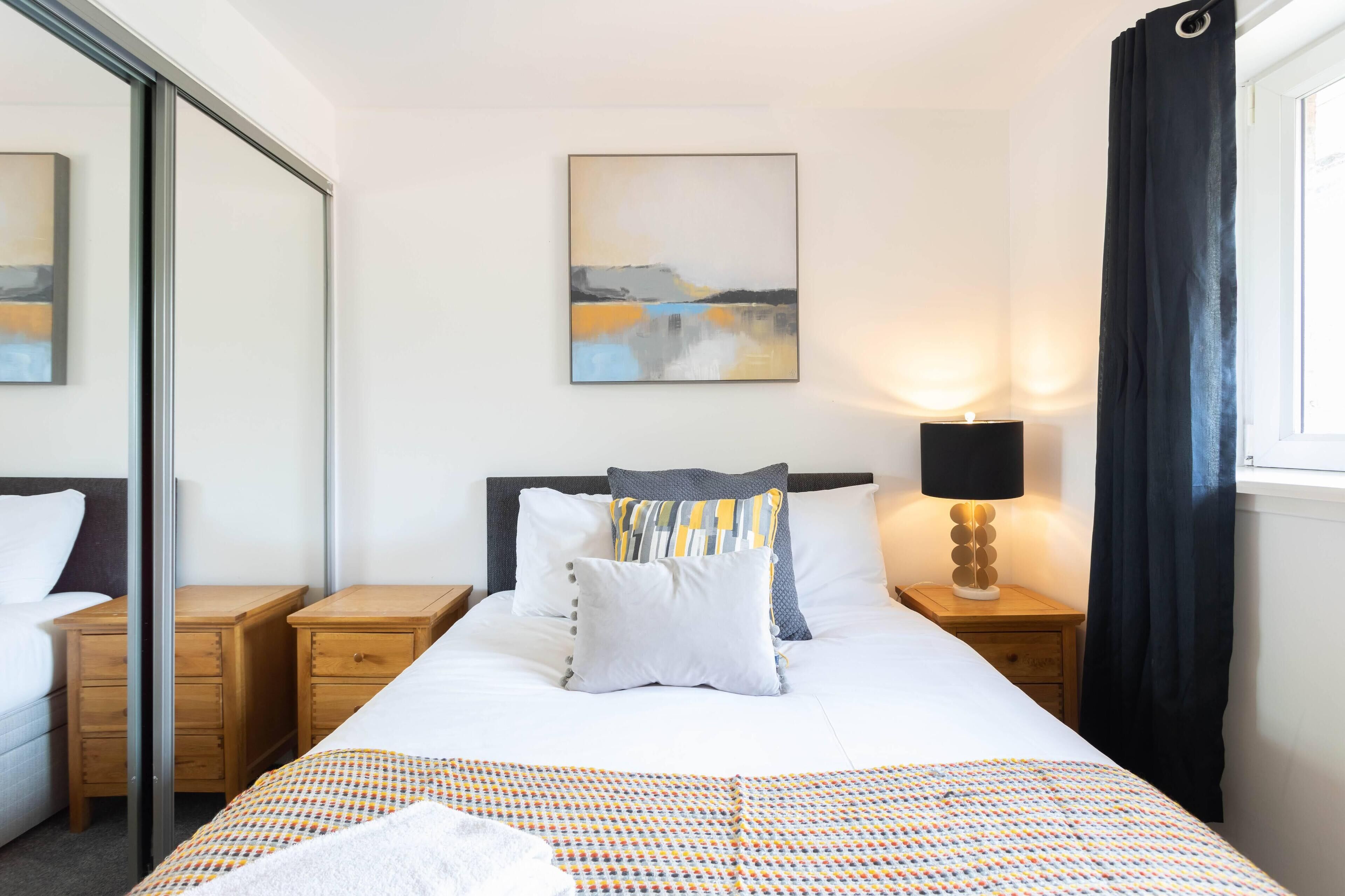 A hotel room with a bed, a nightstand with a lamp, a painting on the wall, and a window with curtains.