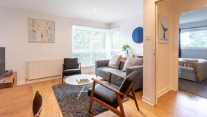 A modern living room with a sofa, chairs, a coffee table, and a TV. There are windows with blinds, a radiator, and a door leading to another room. - Broompark (Glasgow)