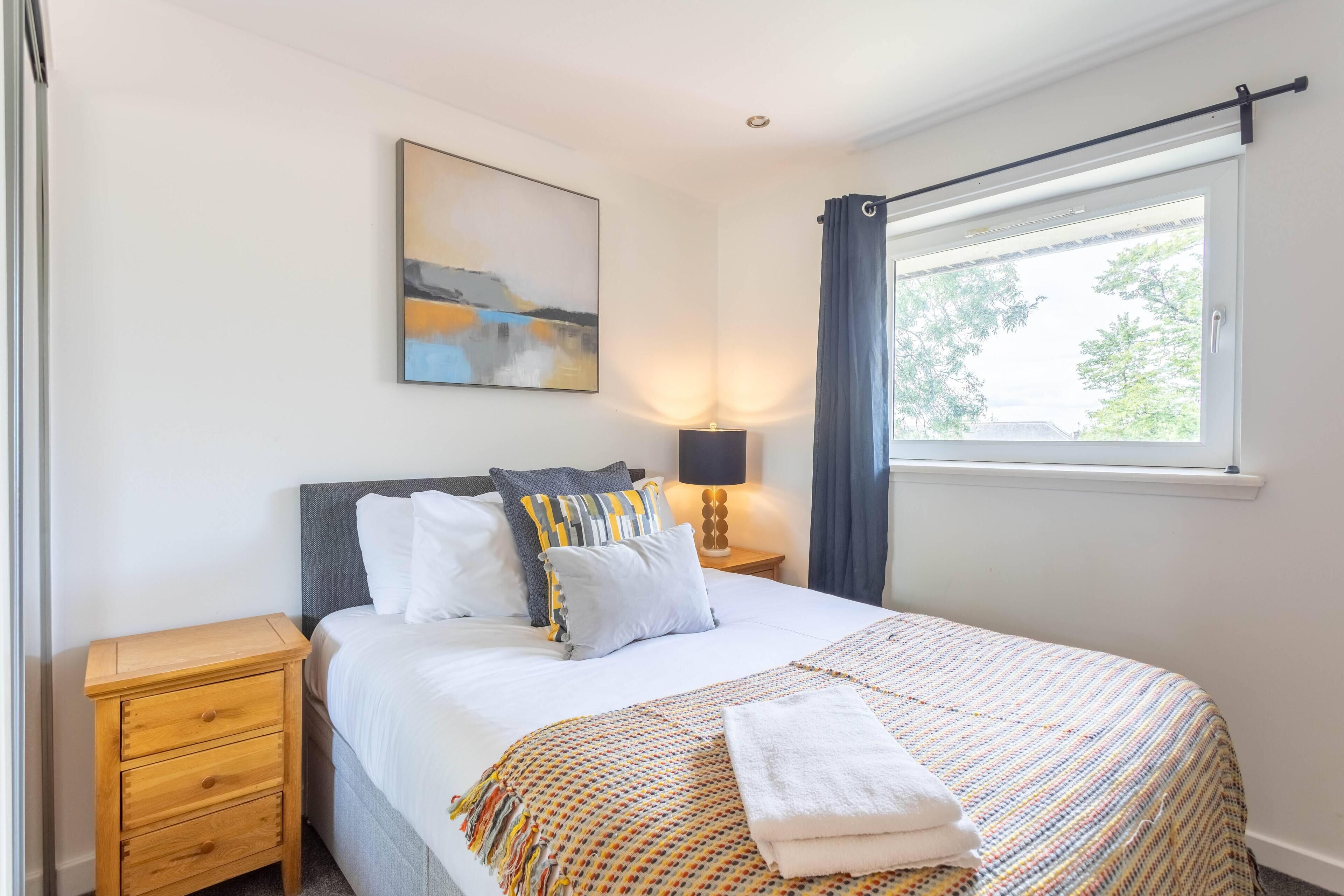 Un dormitorio ordenado con cama, mesita de noche, lámpara y ventana con cortinas.