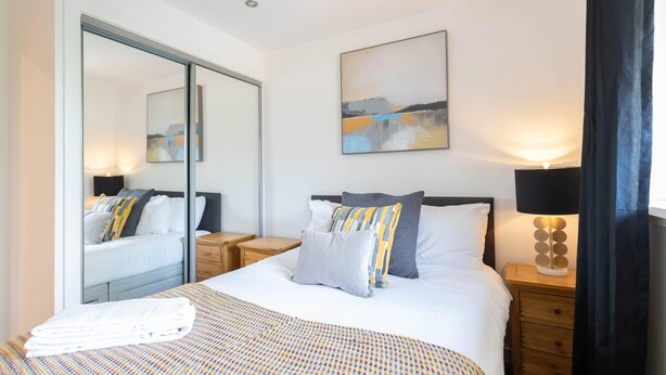 A modern hotel room with a large bed, a bedside table with a lamp, a mirror, and a painting on the wall. - Broompark (Glasgow)