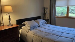 A neatly made bed with a headboard, a bedside table with a lamp, and a window with curtains.