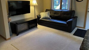 A living room with a flat-screen TV, a dark leather sofa, a lamp, and a window with curtains.