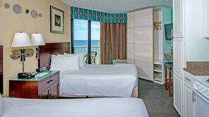 A hotel room with two beds, a desk with a lamp, a TV, and a kitchenette.