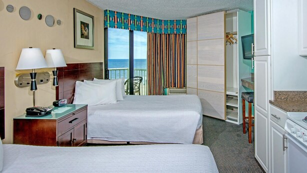 A hotel room with two beds, a desk with a lamp, a TV, and a kitchenette. - The Strand - A Boutique Resort (Myrtle Beach)