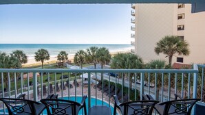 A balcony with a pool and beach view, featuring lounge chairs and a palm tree.
