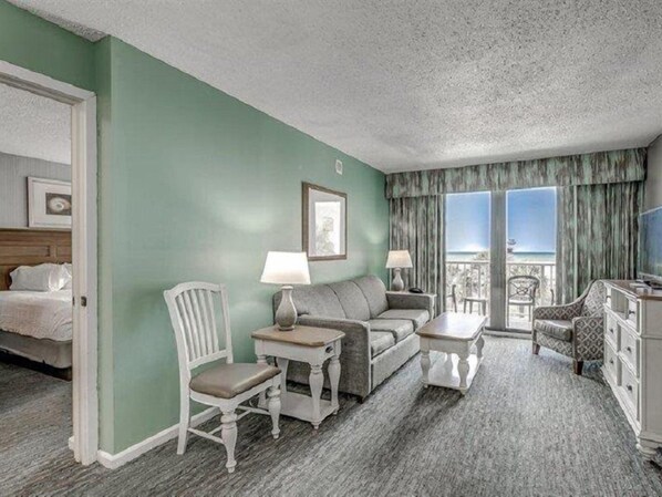 A hotel room with a green wall, a bed, a gray sofa, a white chair, a small table, and a TV. - The Strand - A Boutique Resort (Myrtle Beach)