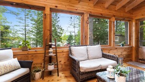 Een houten cabin binnenzicht met een rieten bank, een glazen tafel met planten, en grote ramen met uitzicht op bomen.