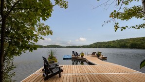 Een houten steiger met lounge stoelen en een klein zwembad, met uitzicht op een kalm meer met heuvels in de verte.