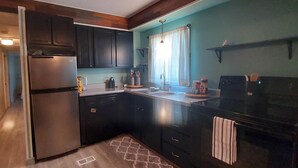 Cocina compacta con armarios oscuros, un refrigerador de acero inoxidable, un lavamanos y una ventana con cortinas.