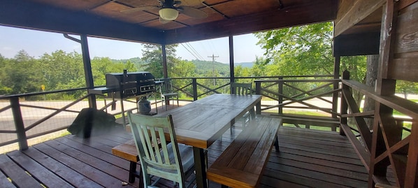 An outdoor covered patio with a wooden table and chairs, a grill, and a view of the surrounding greenery.