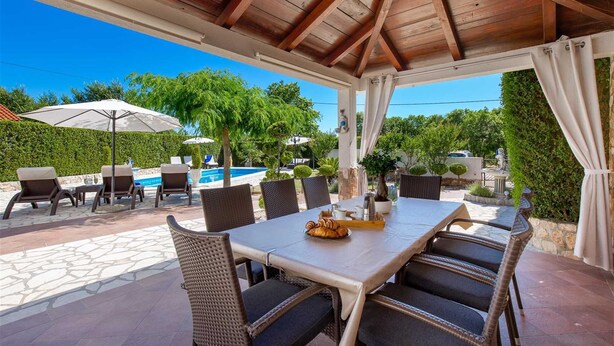 Eine Terrasse im Freien mit einem für zwei Personen gedeckten Tisch, umgeben von Korbstühlen, einem Pool und Grünflächen im Hintergrund.