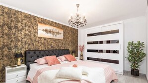 Ein Schlafzimmer mit einer gebogenen Kopfstütze, einem großen Bett mit rosa Bettwäsche, einer Chandelüster, einem Wandbild und einem Blumentopf.