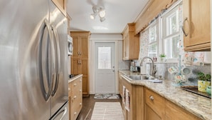 A kitchen with wooden cabinets, stainless steel appliances, a granite countertop, and a window with floral curtains. - The Funky Swan Shack by Lake Morton & Downtown (Lakeland)