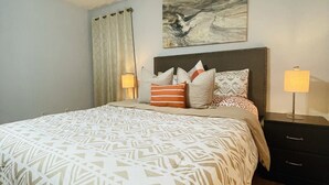 A neatly arranged bedroom with a patterned bedspread, a headboard with multiple pillows, a wall-mounted abstract painting, and bedside lamps.
