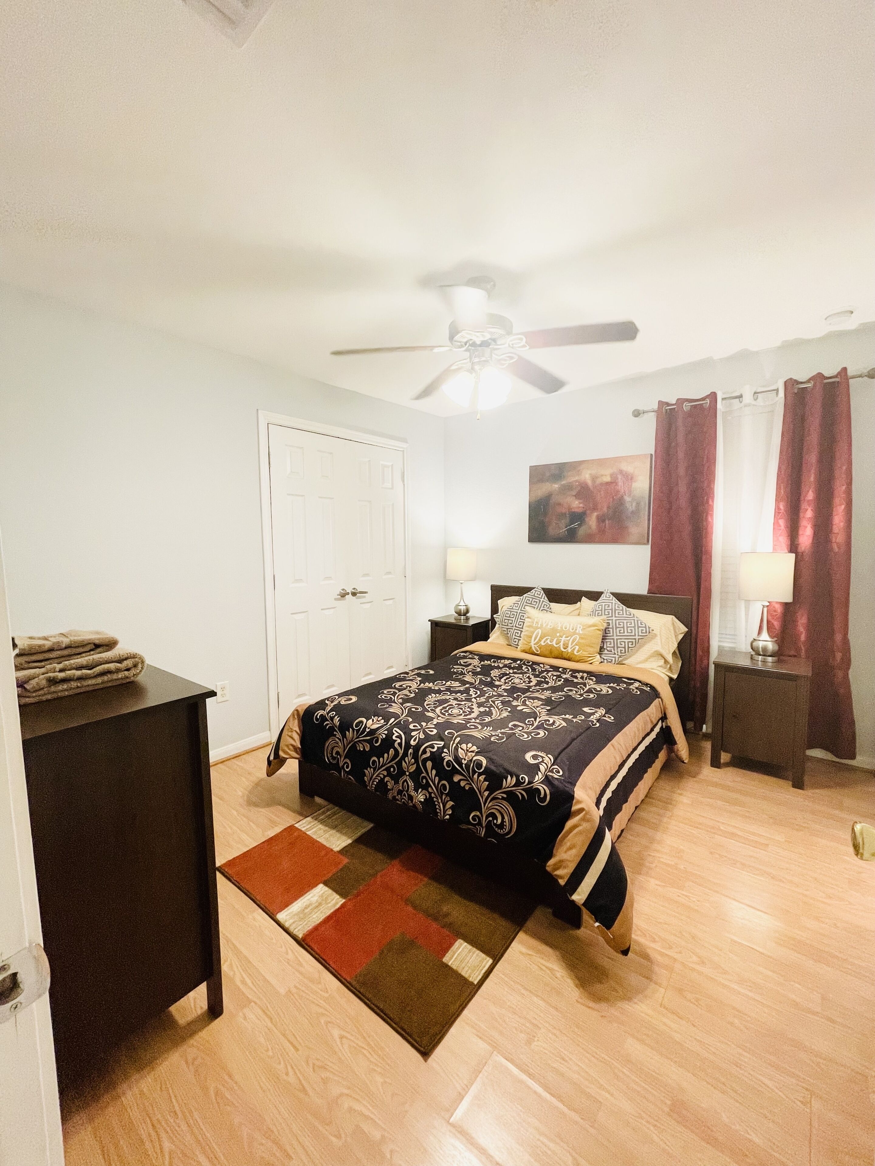 A bedroom with a bed, a ceiling fan, a nightstand, a lamp, a door, and a painting on the wall.
