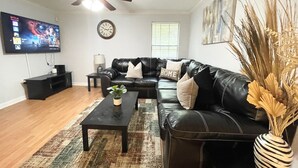 A living room with a black leather sofa, a coffee table, a TV, a clock, and a window with blinds.