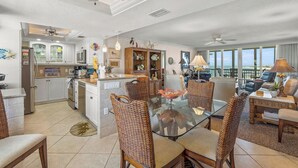 A spacious living area with a glass dining table surrounded by wicker chairs, a kitchen with white cabinets, and a balcony with a view of the ocean. - Crystal Sands Tower Unit 1007 - Beachfront (Sarasota)