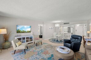 A living room with a TV, sofa, dining table, chairs, and a kitchen area. - Crystal Sands Tower Unit 204 - Beachfront (Sarasota)