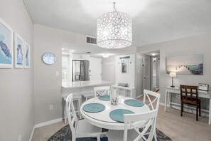 A modern kitchen with white cabinetry, a stainless steel refrigerator, and a dining area with white chairs and a round table. - Crystal Sands Tower Unit 204 - Beachfront (Sarasota)
